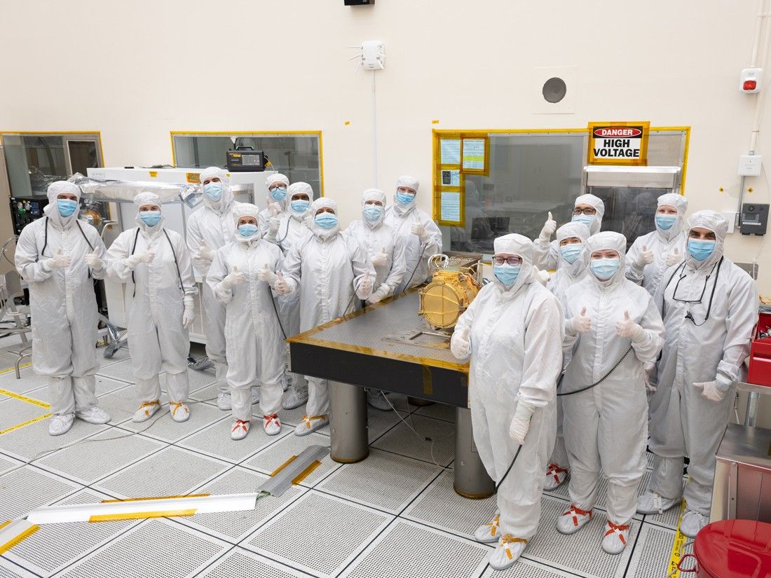 A large group of engineers in full white body coveralls stand surrounding Europa Clipper’s surface dust analyzer. All the engineers are giving a “thumbs up” hand gesture to indicate that the instrument was successfully delivered. Nine engineers stand to the left of a silver workbench, on which the instrument rests, and six stand to the right of the workbench. The instrument is gold colored and about the size of a drum, resting on its side. The instrument is resting upon a silver workbench in the cleanroom.