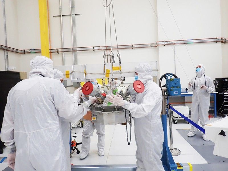 Four engineers wearing white full-body coveralls, gloves, and masks, handle a flat metal structure a few inches thick, to which are attached two telescopes a foot or more long, while a fifth worker in identical attire stands to the side, holding hand-held controls for the crane that’s lofting the hardware.