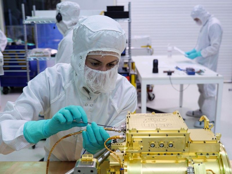 An engineer in white full-body coveralls, a mask, and light blue gloves is shown attaching a wire to the SUDA sensor head. Only the top of the sensor head is visible, and is seen on the lower right portion of the image. The sensor head is gold in color. In the distance, engineers work on other components in the clean room.