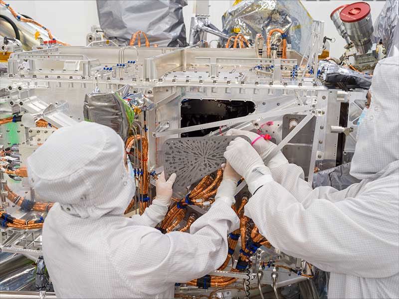 Two workers in white protective clothing hold the triangular-shaped silver-colored vault plate near the Europa Clipper spacecraft.