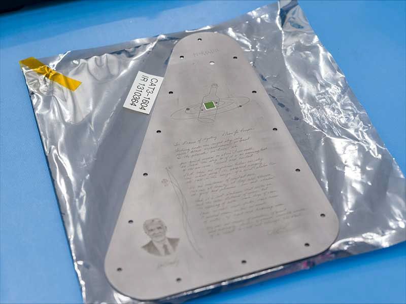 The triangular-shaped silver-colored Europa Clipper Europa Clipper vault plate is covered in a protective plastic bag with a clear front and silver back. There is a bit of gold tape in the upper left corner, and a white label on the bag.