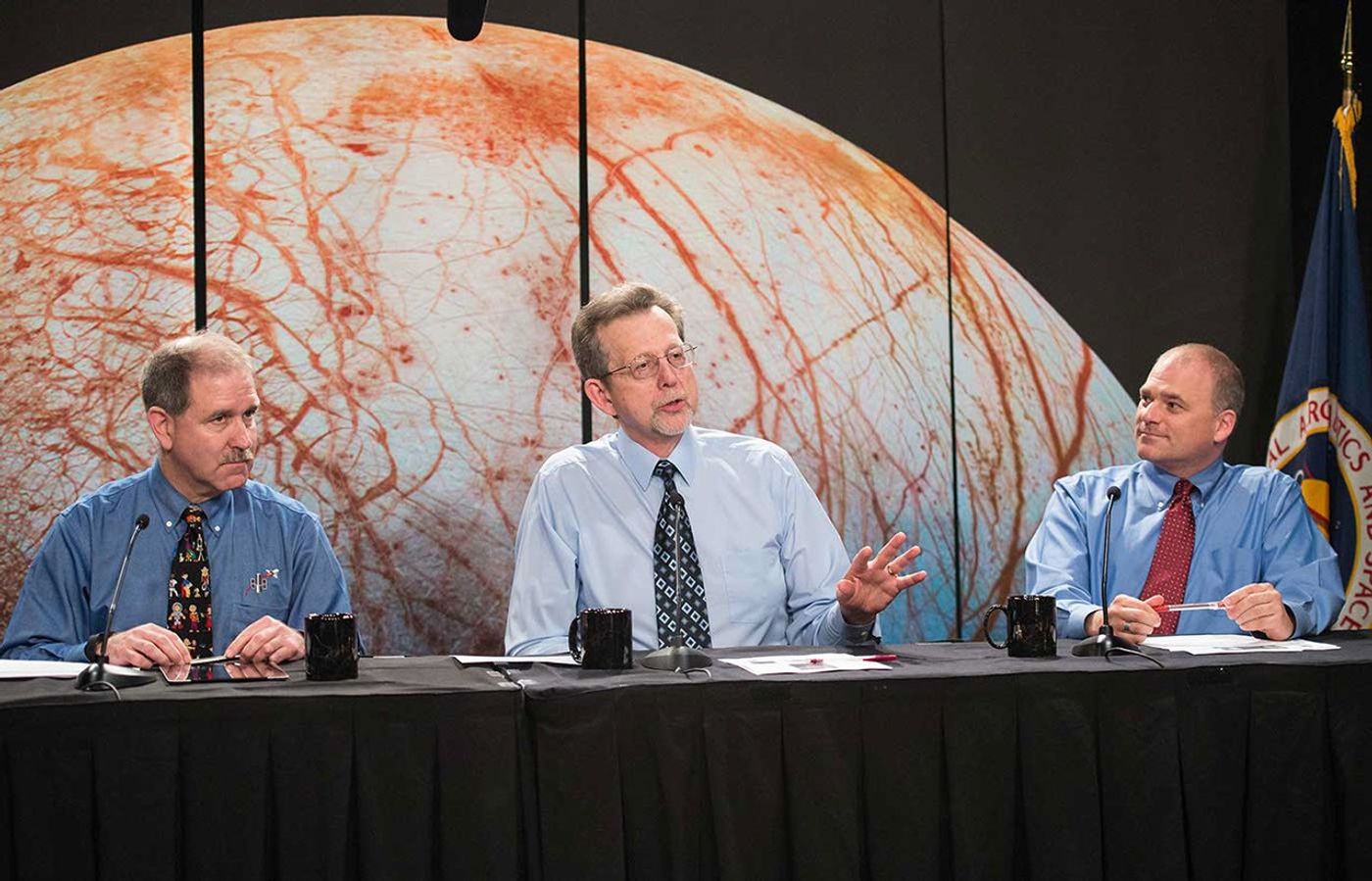 Three men in dress shirts and ties sit at a table with a large image of Europa behind them.