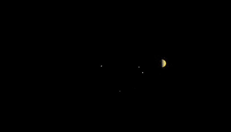 Jupiter appears orange and half lit against the blackness of space. The planet's four largest moons appear nearby as small white dots.
