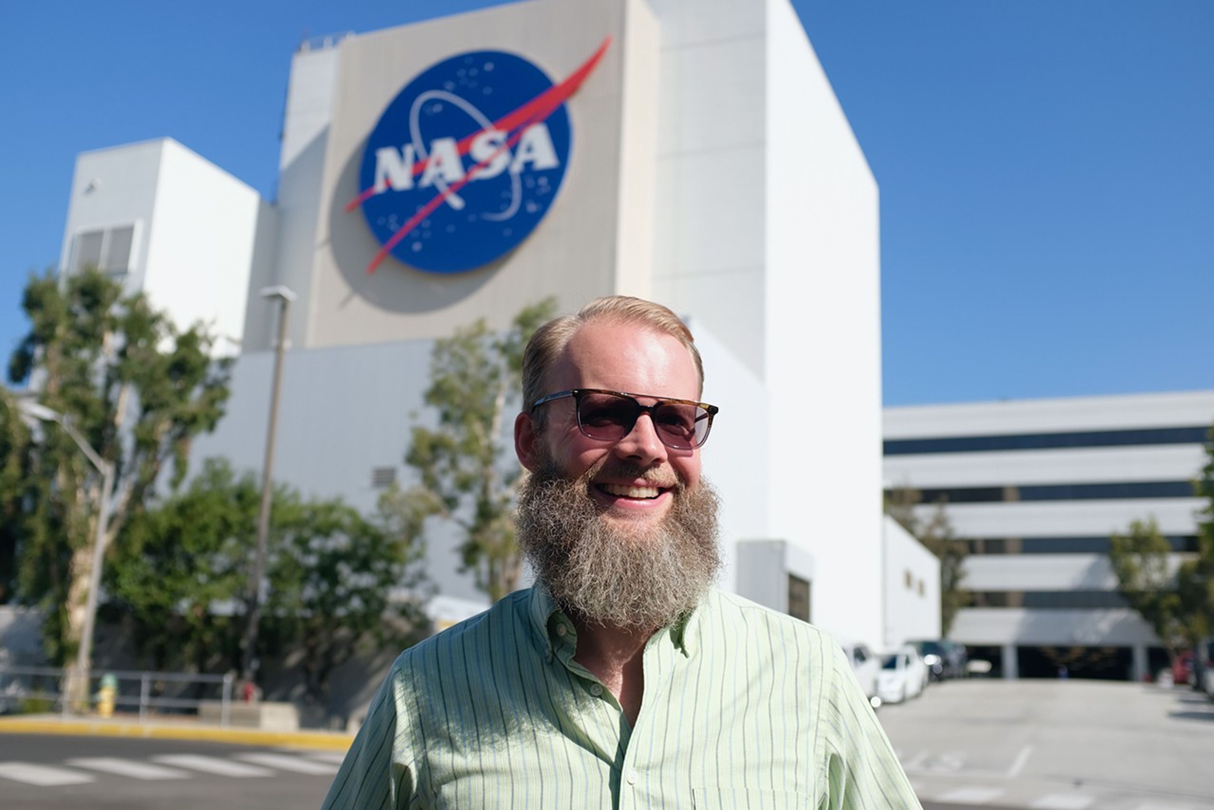 Benjamin Marti, an engineer, stands in the middle of the image wearing a green stripped shirt and brown sunglasses. Behind the engineer, you cans a multistory building with the NASA logo on it.