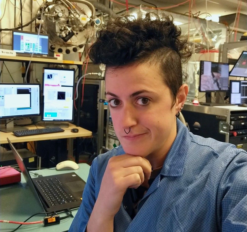 An engineer with a blue button up shirt poses for a photo in lab, with screens and other electrical equipment seen behind them.