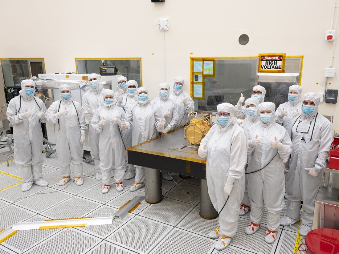 A large group of engineers in full white body coveralls stand surrounding Europa Clipper’s surface dust analyzer. All the engineers are giving a “thumbs up” hand gesture to indicate that the instrument was successfully delivered. Nine engineers stand to the left of a silver workbench, on which the instrument rests, and six stand to the right of the workbench. The instrument is gold colored and about the size of a drum, resting on its side. The instrument is resting upon a silver workbench in the cleanroom.