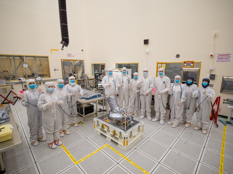 Twelve engineers and technicians dressed in white protective “bunny suits” stand behind and beside to Europa Clipper’s narrow-angle camera (NAC). The NAC is on a platform with rods sticking straight up at the corners of the platform. It’s wrapped in silver-colored insulating material, it has a round base about the size of a tire. The lens is sticking out at an angle and is laced up inside the insulating material. Yellow tape on the floor marks the area around NAC. Equipment and gear sits on tables behind, and to the sides of the workers.