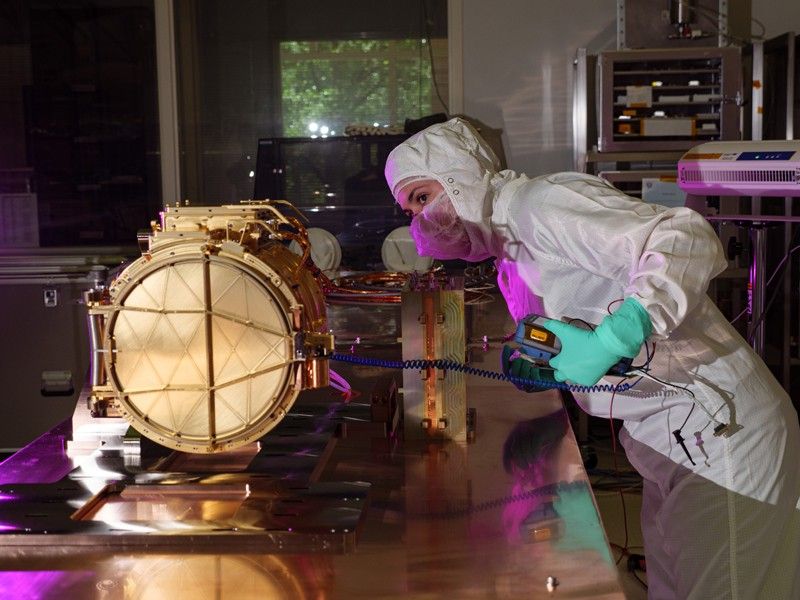 The SUDA sensor head rests upon a table in a clean room. The sensor head is gold in color, shaped slightly like a drum on its side with a door facing the viewer that is closed. An engineer in white coveralls, a mask, and light blue gloves is on the right side of the image and is seen peering over the instrument. The engineer is holding a handheld device attached a blue wire, which itself is attached to the sensor head, and they perform a test. Is a purple light is shown on the table and on the front of the engineer.