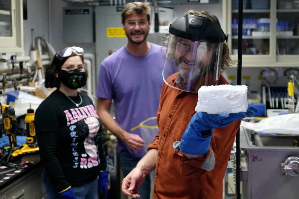 Inside the Ocean Worlds Lab, Dr. Kevin Peter Hand displays a brick-sized block of ice while wearing a blue protective glove and face shield. With Hand are Sarah N. Yearicks, mechanical engineer and lead of JPL’s Extraterrestrial Simulants Lab, and Jeff Foster, technologist at JPL’s Ocean Worlds Lab. To the right of the image is a portion of the Ark, a gray vacuum chamber about the size of a deep freezer. In the background are various tools, instruments, and cabinets.