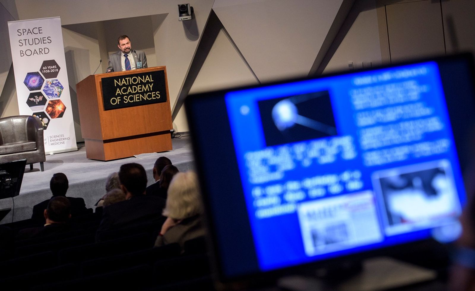 In this image, Alexander Moiseev stands at a wooden podium labeled "National Academy of Sciences," delivering a presentation to a seated audience. To his left, a vertical banner for the "Space Studies Board" commemorates "60 Years 1958–2018," featuring various celestial and satellite images. In the blurred foreground, a monitor displays a blue presentation slide with text and a small image of the Sputnik satellite, capturing a moment from the symposium's historical retrospective.