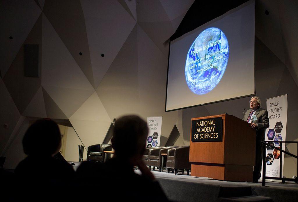 A wide shot from the perspective of the stage at the National Academy of Sciences shows Michael Freilich, Director of NASA's Earth Science Division, addressing a large audience from a wooden podium. Above him, a large projector screen displays a brilliant image of Earth from space with the title "Follow the Water: Monitoring and Understanding Earth's Hydrologic Cycle From Space". To the right of the stage, a "Space Studies Board" banner commemorates 60 years of scientific achievement (1958–2018), while the darkened auditorium is filled with attendees listening to the presentation.