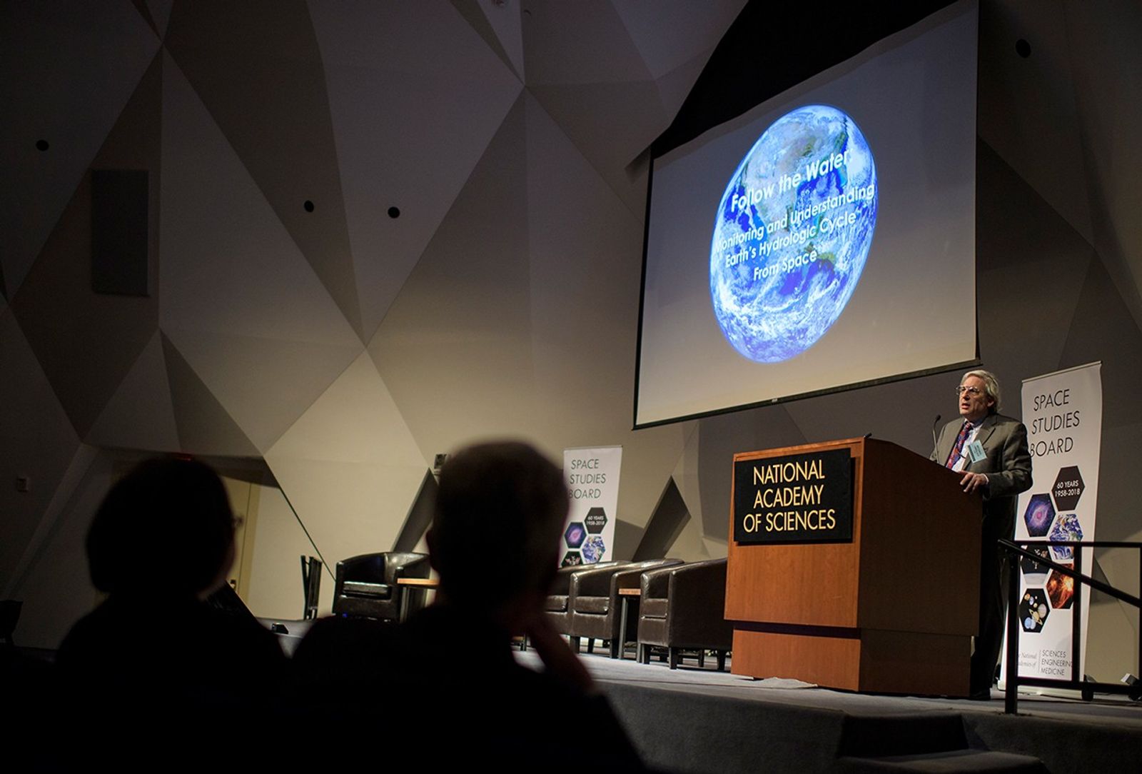 A wide shot from the perspective of the stage at the National Academy of Sciences shows Michael Freilich, Director of NASA's Earth Science Division, addressing a large audience from a wooden podium. Above him, a large projector screen displays a brilliant image of Earth from space with the title "Follow the Water: Monitoring and Understanding Earth's Hydrologic Cycle From Space". To the right of the stage, a "Space Studies Board" banner commemorates 60 years of scientific achievement (1958–2018), while the darkened auditorium is filled with attendees listening to the presentation.