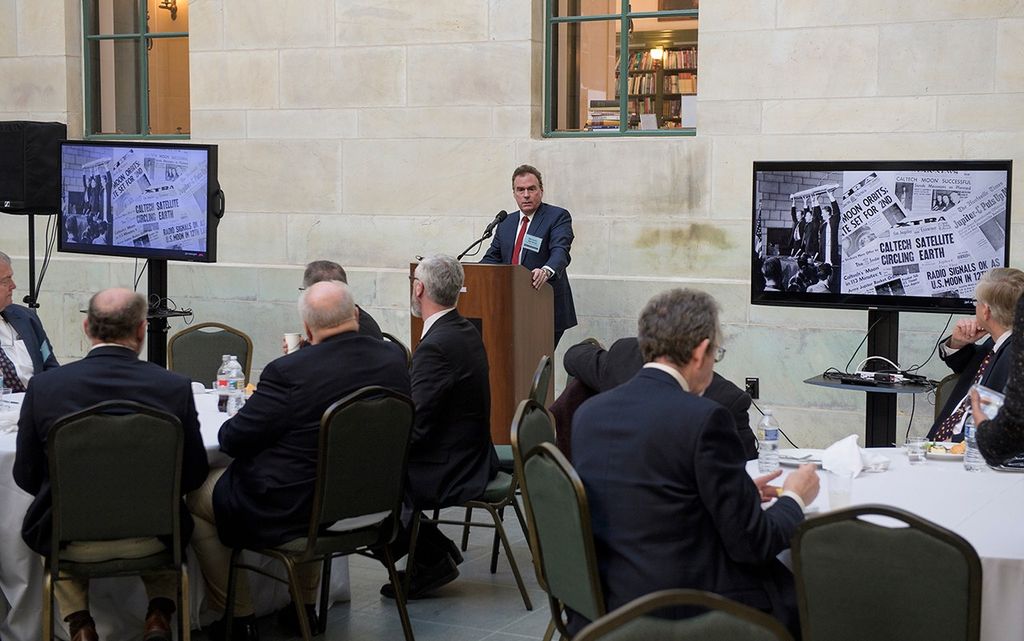 A man in a dark suit and red tie speaks at a wooden podium during a luncheon in a room with light stone walls. He is flanked by two large monitors displaying a collage of historic newspaper headlines and the iconic photo of the Explorer 1 satellite being held aloft. In the foreground, several guests are seated at round tables covered in white cloths, their backs to the camera as they listen to the presentation.