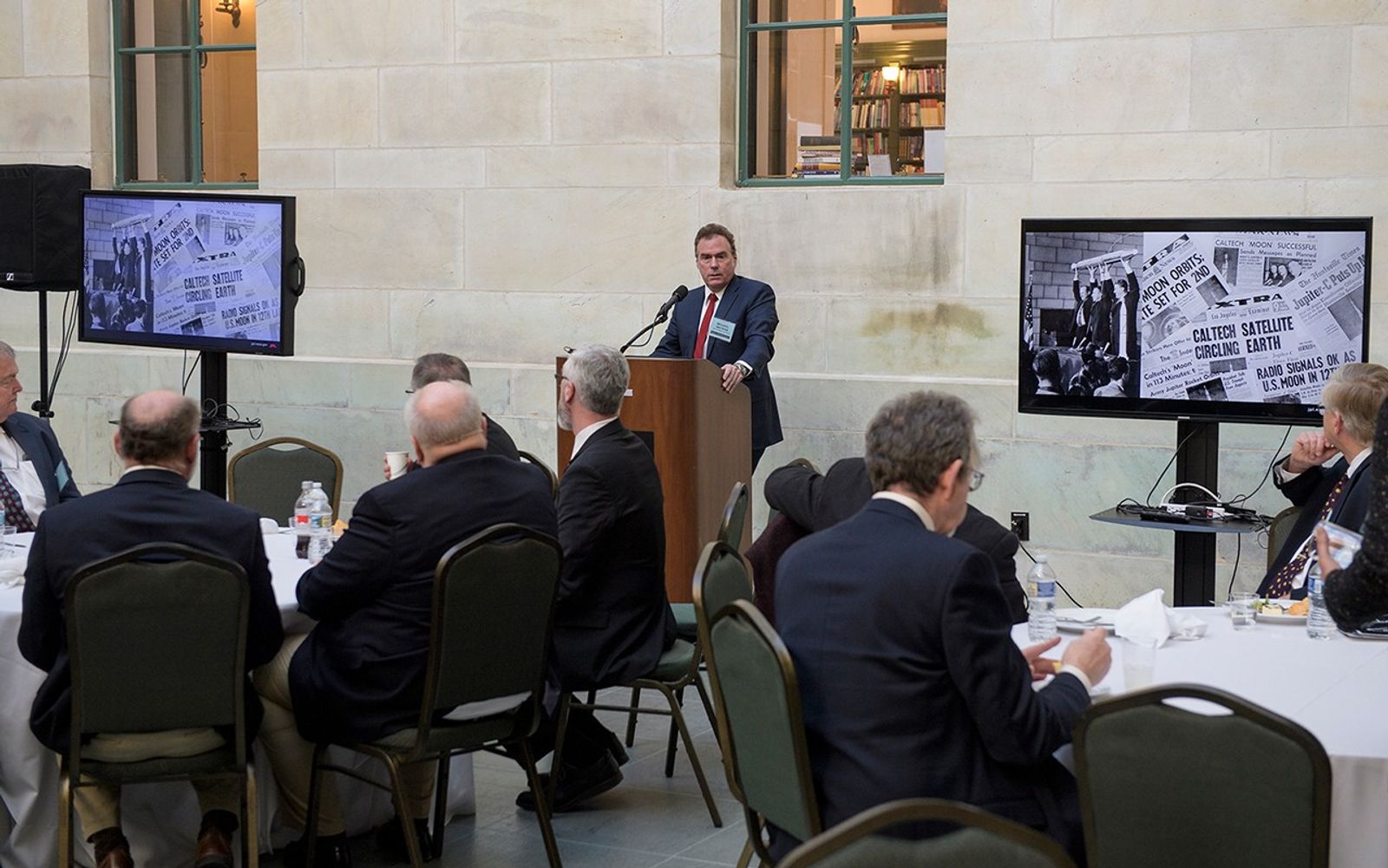 A man in a dark suit and red tie speaks at a wooden podium during a luncheon in a room with light stone walls. He is flanked by two large monitors displaying a collage of historic newspaper headlines and the iconic photo of the Explorer 1 satellite being held aloft. In the foreground, several guests are seated at round tables covered in white cloths, their backs to the camera as they listen to the presentation.