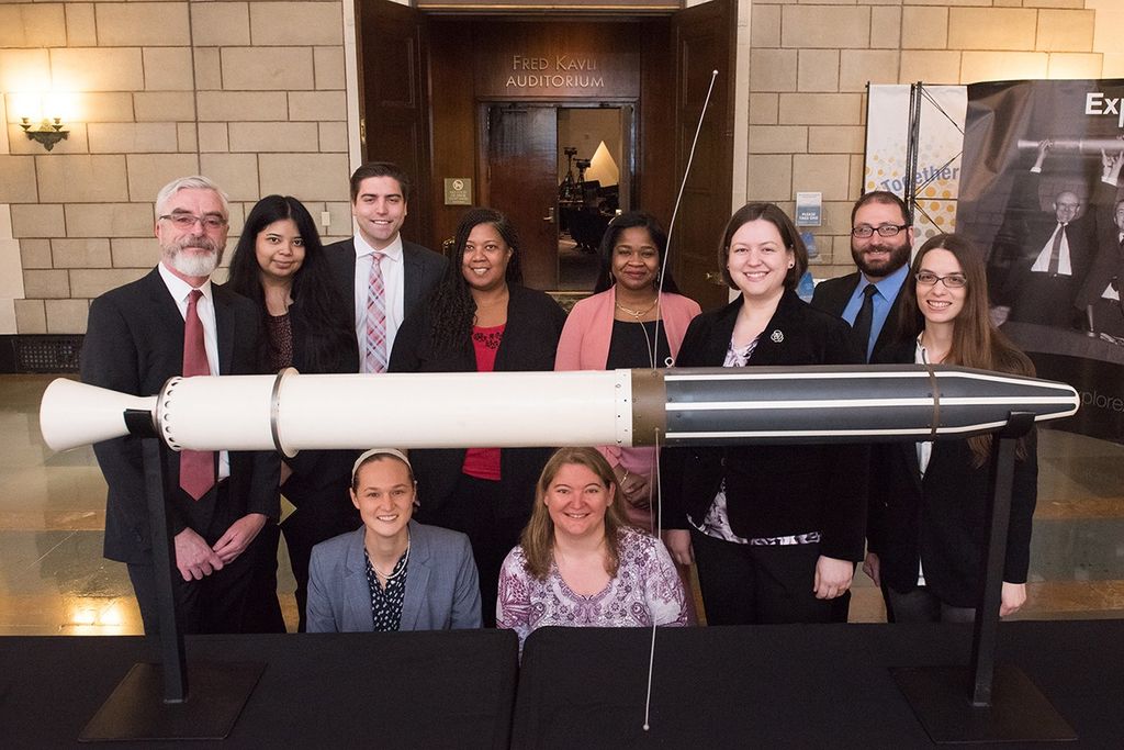 A diverse group of ten people stands and kneels behind a full-scale model of the Explorer 1 satellite, posed in front of the entrance to the Fred Kavli Auditorium. The group is dressed in professional attire, with two women kneeling in the front and eight men and women standing behind them in a grand stone hall. The white and black satellite model, featuring its signature whip antennas, rests on two black stands in the foreground, while a banner depicting the historic "lift" of the original satellite is visible to the right.