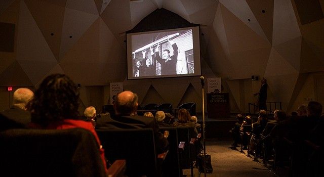 A view from the back of a darkened, geometric-walled auditorium shows an audience watching a large projector screen. The screen displays a historic black-and-white photograph of three men triumphantly holding a model of the Explorer 1 satellite above their heads. To the right of the screen, a speaker stands near a podium labeled "National Academy of Sciences," with signs for the "Space Studies Board" visible on the stage.