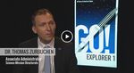 A medium close-up shot features Dr. Thomas Zurbuchen, NASA’s Associate Administrator for the Science Mission Directorate, speaking during a video interview. He is wearing a dark suit with a small NASA pin and a blue-and-white patterned tie. In the background, a brightly lit monitor displays a graphic poster that reads "GO! EXPLORER 1" in large blue and black letters, alongside a stylized illustration of the satellite launching into a starry sky to mark "60 Years of America in Space".