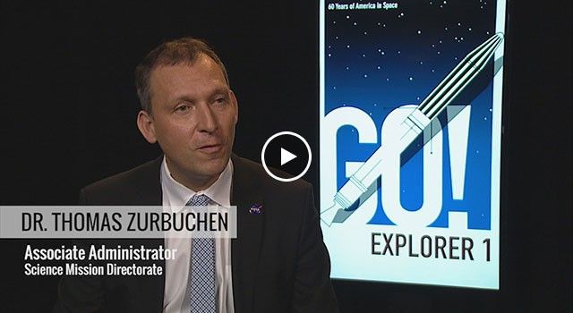 A medium close-up shot features Dr. Thomas Zurbuchen, NASA’s Associate Administrator for the Science Mission Directorate, speaking during a video interview. He is wearing a dark suit with a small NASA pin and a blue-and-white patterned tie. In the background, a brightly lit monitor displays a graphic poster that reads "GO! EXPLORER 1" in large blue and black letters, alongside a stylized illustration of the satellite launching into a starry sky to mark "60 Years of America in Space".