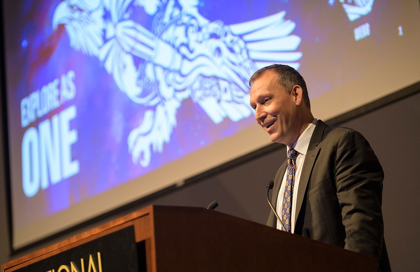 A low-angle, medium shot features Thomas Zurbuchen standing at a wooden podium, smiling as he looks toward an audience. He is wearing a dark suit and a distinctive blue tie with a bright orange and yellow pattern. Behind him, a large screen displays a vibrant purple and blue graphic with the bold white text "EXPLORE AS ONE" and a stylized illustration of an eagle carrying a rocket.
