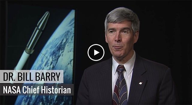 A medium close-up shot features Dr. Bill Barry, NASA’s Chief Historian, speaking during a video interview. He is dressed in a dark pinstripe suit with a small NASA pin and a patterned tie. In the background, a large monitor displays a dramatic image of the Explorer 1 satellite in orbit against the curve of the Earth, highlighting the historical significance of the mission he is discussing.