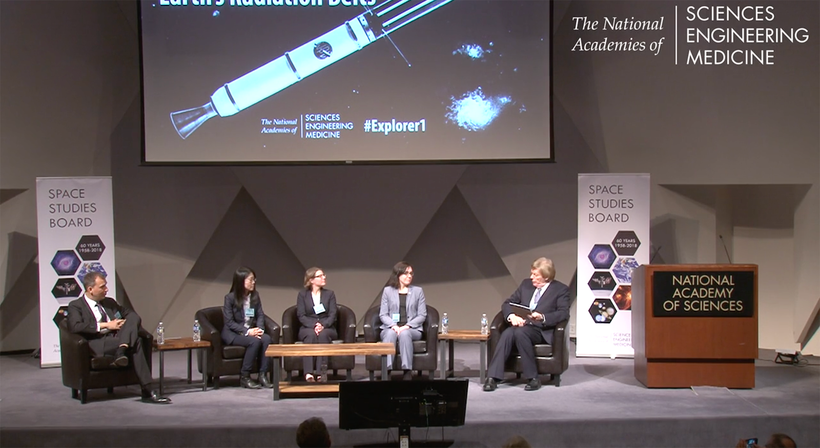 In this wide shot of a stage at the National Academy of Sciences, five panelists are seated in leather armchairs for a discussion on the future of space science. Above them, a large screen displays a diagram of the Explorer 1 satellite over the hashtag "#Explorer1" and the logos for the National Academies of Sciences, Engineering, and Medicine. To the left and right of the stage, "Space Studies Board" banners mark the 60th anniversary of the mission. The panel is positioned in front of a modern, geometric wall, with a wooden podium labeled "National Academy of Sciences" standing to the far right.