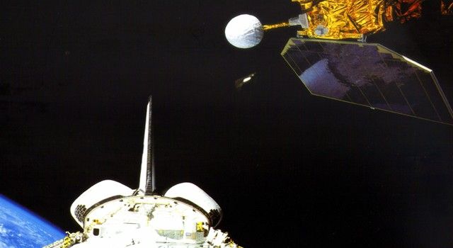 An action shot from space shows the Earth Radiation Budget Satellite (ERBS) being deployed from the cargo bay of a Space Shuttle. In the upper right, the ERBS satellite is visible, covered in gold foil with a large, dark rectangular solar panel extended and a white spherical instrument protruding from its side. Below it, the white tail fin and open cargo bay doors of the Space Shuttle are prominent against the pitch-black void of space. In the bottom left corner, the curved limb of the blue Earth with white clouds is visible, providing a sense of scale and orbit.