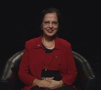 A medium-shot portrait of a woman with dark hair and glasses, sitting in a black leather armchair against a solid black background. She is wearing a vibrant red blazer over a black top, accessorized with a gold necklace and a thin lanyard. She has a warm smile and her hands are calmly clasped in her lap. The lighting is focused on her, creating a professional and formal atmosphere.
