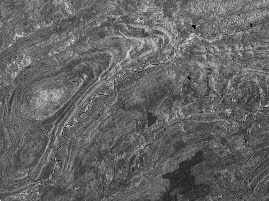 A grayscale satellite image shows a vast, textured landscape from a high-altitude top-down perspective. The terrain is characterized by prominent, winding geological folds and ridges that create a series of concentric, wavy patterns across the surface. A dark, thin river or stream meanders through the center of the image, cutting across the rugged topography. The varied shades of gray indicate different types of land cover or rock formations, with some darker patches likely representing water or dense vegetation and lighter areas highlighting the crests of the ridges.