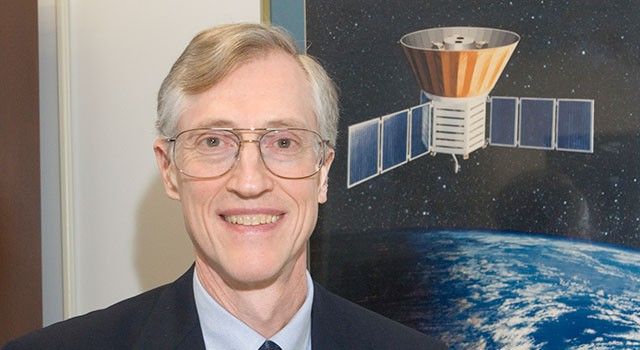 A head-and-shoulders portrait of a smiling man with fair skin, light brown hair swept to the side, and gold-rimmed glasses. He is wearing a dark suit jacket over a light blue dress shirt and a dark tie. Behind him is a poster or illustration depicting a satellite with solar panels orbiting Earth against a dark, starry space background. The lighting is bright and professional, suggesting an office or academic setting.