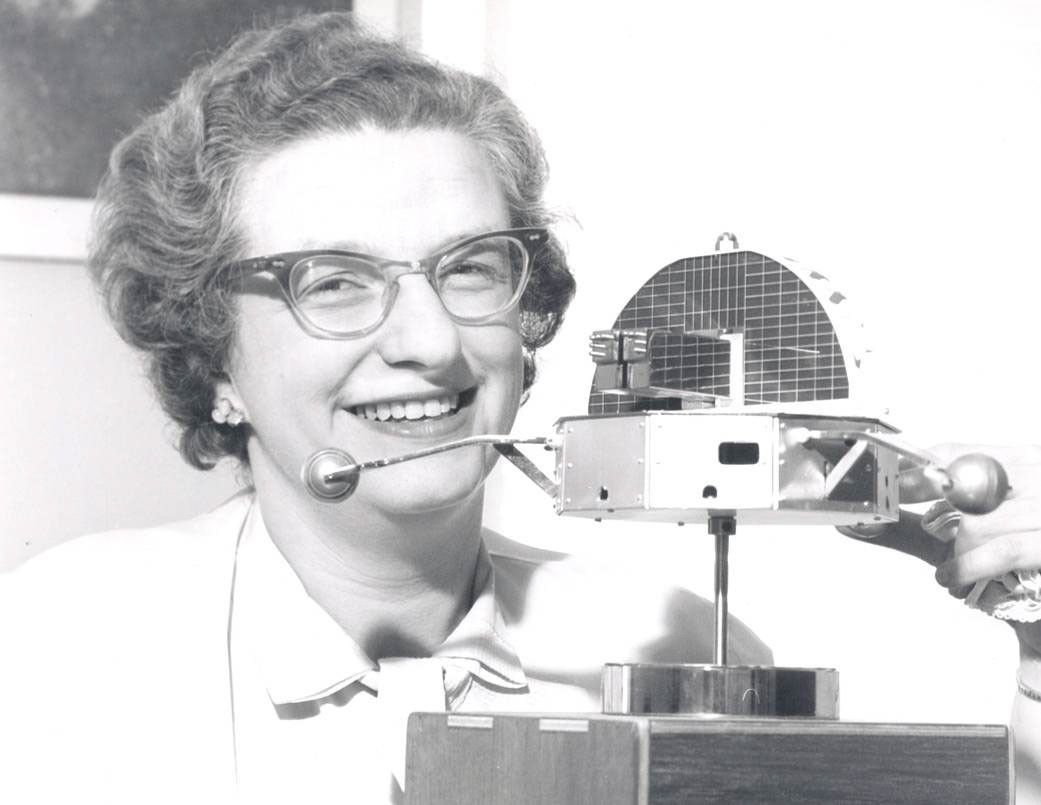 A black-and-white historical photograph shows a smiling woman with short, wavy hair and cat-eye glasses looking toward the camera. She is positioned next to a technical model of a satellite, which features a grid-patterned dish and protruding scientific instruments. The woman is wearing a light-colored button-down shirt, and the background is a simple, bright indoor space, highlighting her and the model as the primary subjects.