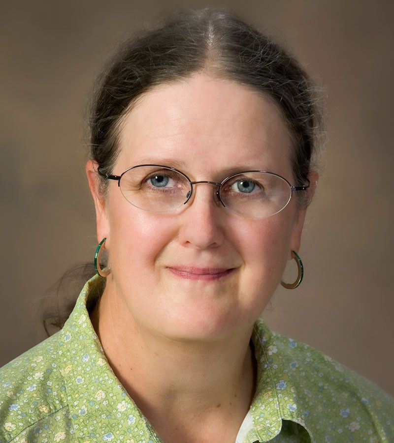 A close-up, professional headshot of a woman with fair skin, blue eyes, and light brown hair pulled back from her forehead. She is wearing thin, oval-shaped glasses and green hoop earrings that match her light green, floral-patterned button-down shirt. She has a gentle, closed-mouth smile and is set against a soft, neutral brown gradient background.
