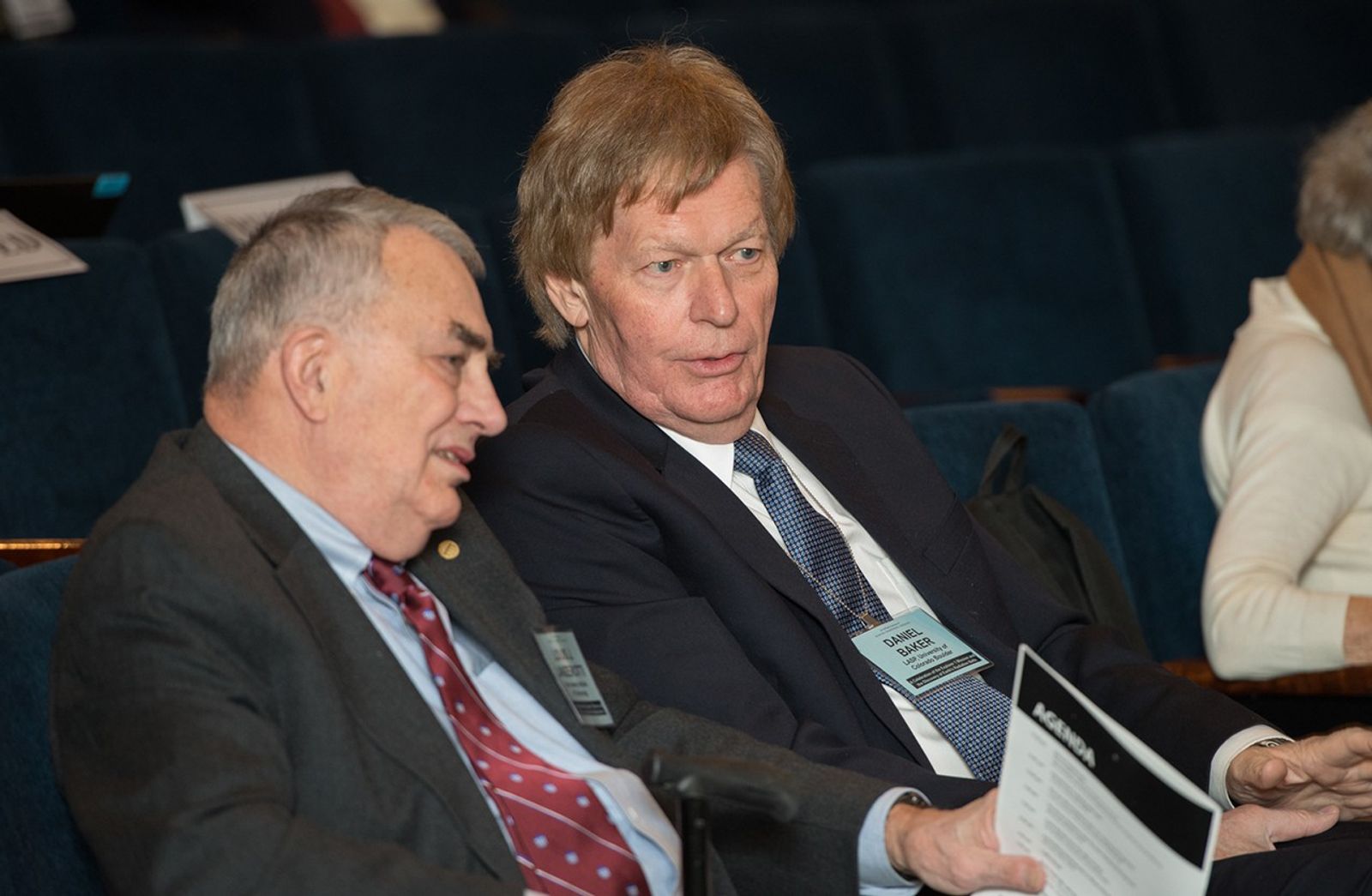 In this close-up shot, two distinguished men are seen in deep conversation while seated in an auditorium. Louis Lanzerotti (left), a research professor at the New Jersey Institute of Technology, and Daniel Baker (right), director of the Laboratory for Atmospheric and Space Physics at the University of Colorado Boulder, are attending an event at the National Academy of Sciences. Both men wear professional suits and identification badges, with Baker holding an event agenda as they talk. The background shows the dark blue seats of the auditorium, where they are participating in a symposium celebrating the 60th anniversary of the Explorer 1 mission.
