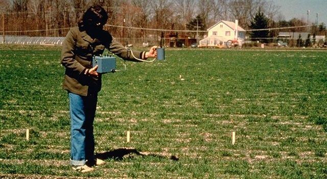 A wide outdoor shot shows a person standing in a grassy field, holding a scientific instrument used for field measurements. The individual is wearing a dark olive jacket and blue jeans, with their face partially obscured by a shadow. They hold a blue rectangular device in one hand, connected by a cable to a handheld cylindrical sensor pointed toward the ground. In the background, a residential house and a line of bare trees are visible under a clear sky.
