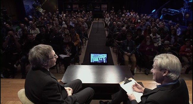 A high-angle view from a stage shows two men in suits seated and facing each other in conversation, with one holding a clipboard. They are positioned in the foreground, overlooking a large, seated audience in a dimly lit auditorium. A small monitor displaying a video feed sits on a table between the speakers, while a long center aisle divides the crowd, leading toward the back of the room where various scientific displays are visible.