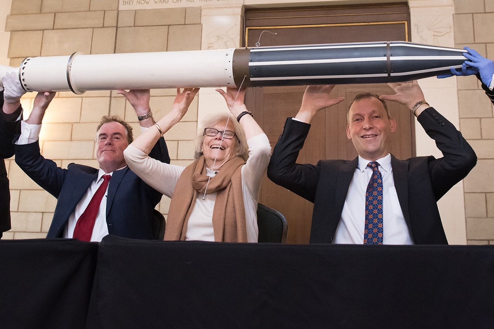 Three people—two men in suits and a woman in a tan sweater and scarf—recreate a famous historic pose by holding a full-scale model of the Explorer 1 satellite above their heads. They are seated behind a black-clothed table in a grand hall with stone walls and a tall wooden door. The woman in the center smiles broadly as they support the white and black cylindrical model, which features thin wire antennas and is being steadied by gloved hands at either end.