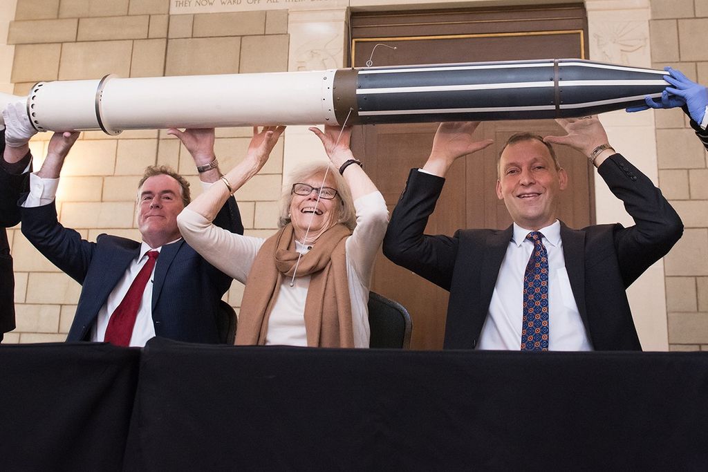 Three people—two men in suits and a woman in a tan sweater and scarf—recreate a famous historic pose by holding a full-scale model of the Explorer 1 satellite above their heads. They are seated behind a black-clothed table in a grand hall with stone walls and a tall wooden door. The woman in the center smiles broadly as they support the white and black cylindrical model, which features thin wire antennas and is being steadied by gloved hands at either end.