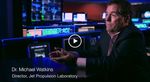 In this medium close-up shot, Dr. Michael Watkins, the former director of NASA's Jet Propulsion Laboratory, is featured during an interview reflecting on the legacy of Explorer 1. He is dressed in a dark suit and seated at a reflective black table, gesturing with his hands as he speaks. The background of the Space Flight Operations Facility (SFOF) is visible, filled with glowing blue monitors, a desk labeled "VOYAGER ACE," and various mission control data displays that symbolize the decades of space exploration that followed the first U.S. satellite.
