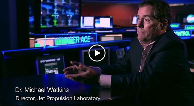 In this medium close-up shot, Dr. Michael Watkins, the former director of NASA's Jet Propulsion Laboratory, is featured during an interview reflecting on the legacy of Explorer 1. He is dressed in a dark suit and seated at a reflective black table, gesturing with his hands as he speaks. The background of the Space Flight Operations Facility (SFOF) is visible, filled with glowing blue monitors, a desk labeled "VOYAGER ACE," and various mission control data displays that symbolize the decades of space exploration that followed the first U.S. satellite.