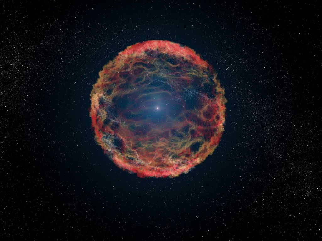 A small white sphere glows at the center of a colorful, spherical-shaped shell. Most of the outer edges are red and yellow, with trails of red, yellow, green, and blue dust inside the shell. This is against the black backdrop of space with countless stars scattered throughout.