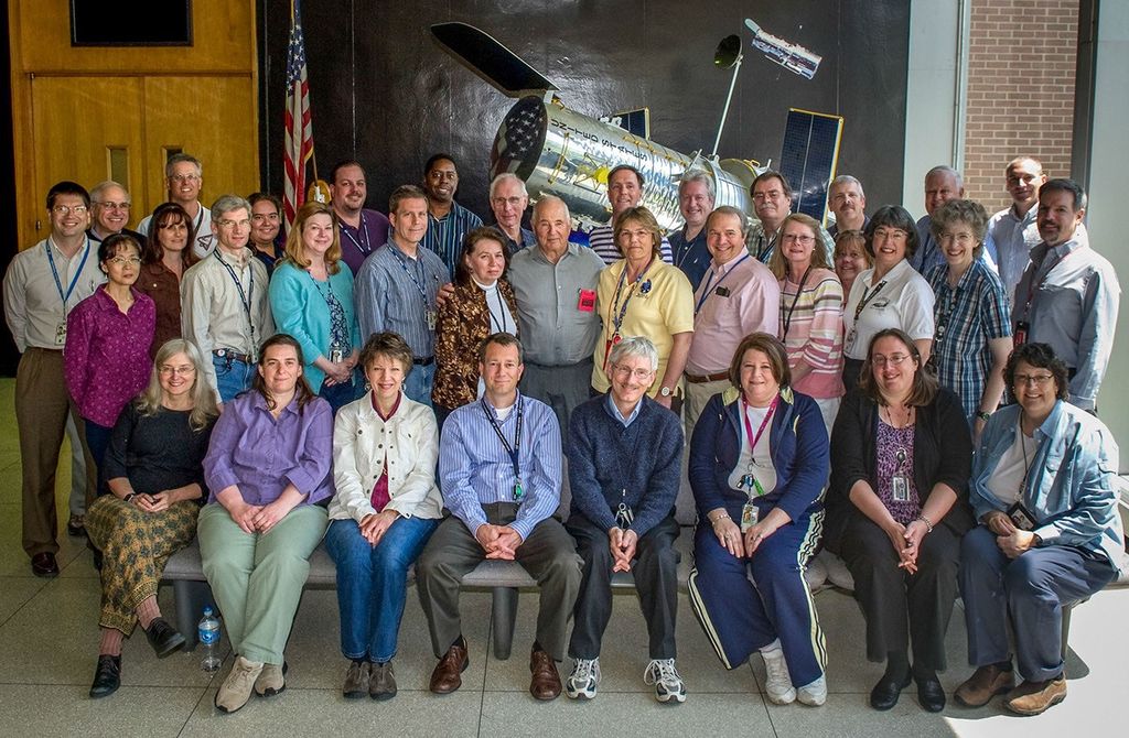 Astronaut Bill Anders stands in the center of a group of 35 people in 3 rows, standing in front of a 1/5 scale model of Hubble.