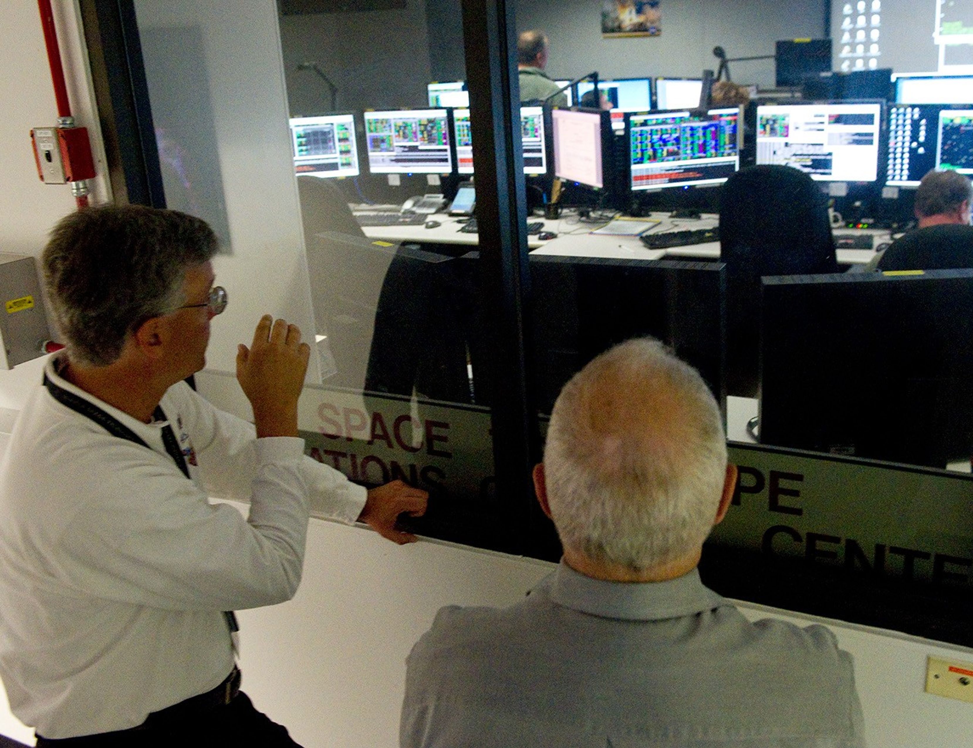 Jim Jeletic, left, and Bill Anders, right, face away from the camera through the glass window into the Mission Operations Room. In the room, three people can be seen sitting in front of rows of computers.