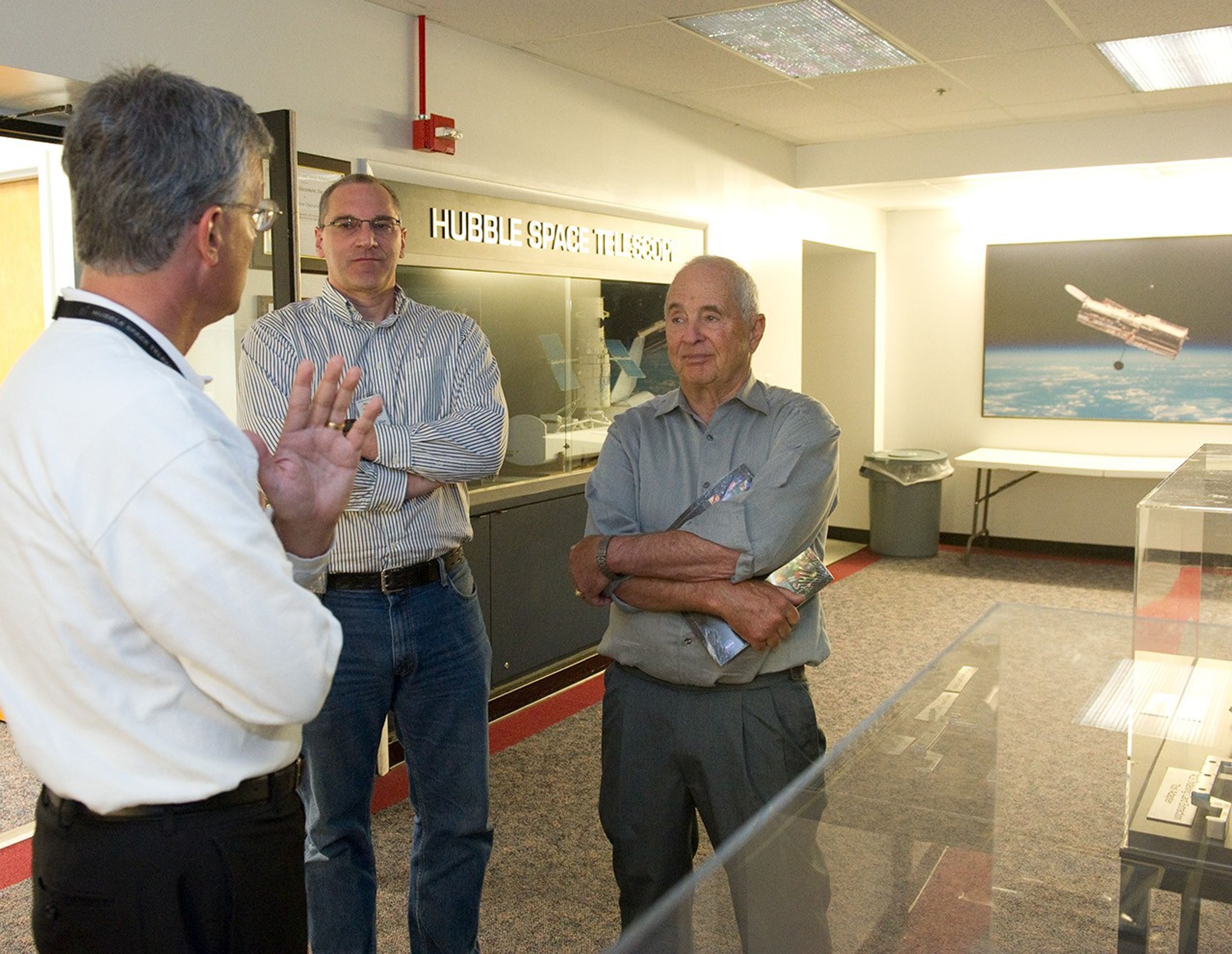 Jim Rice and Bill Anders stand near the Hubble tool cases, facing towards the camera, on the left and right, respectively. Jim Jeletic stands closer to the camera, facing towards them and talking. Behind them, a Hubble display case is visible, along with a wall photo of Hubble over the Earth.