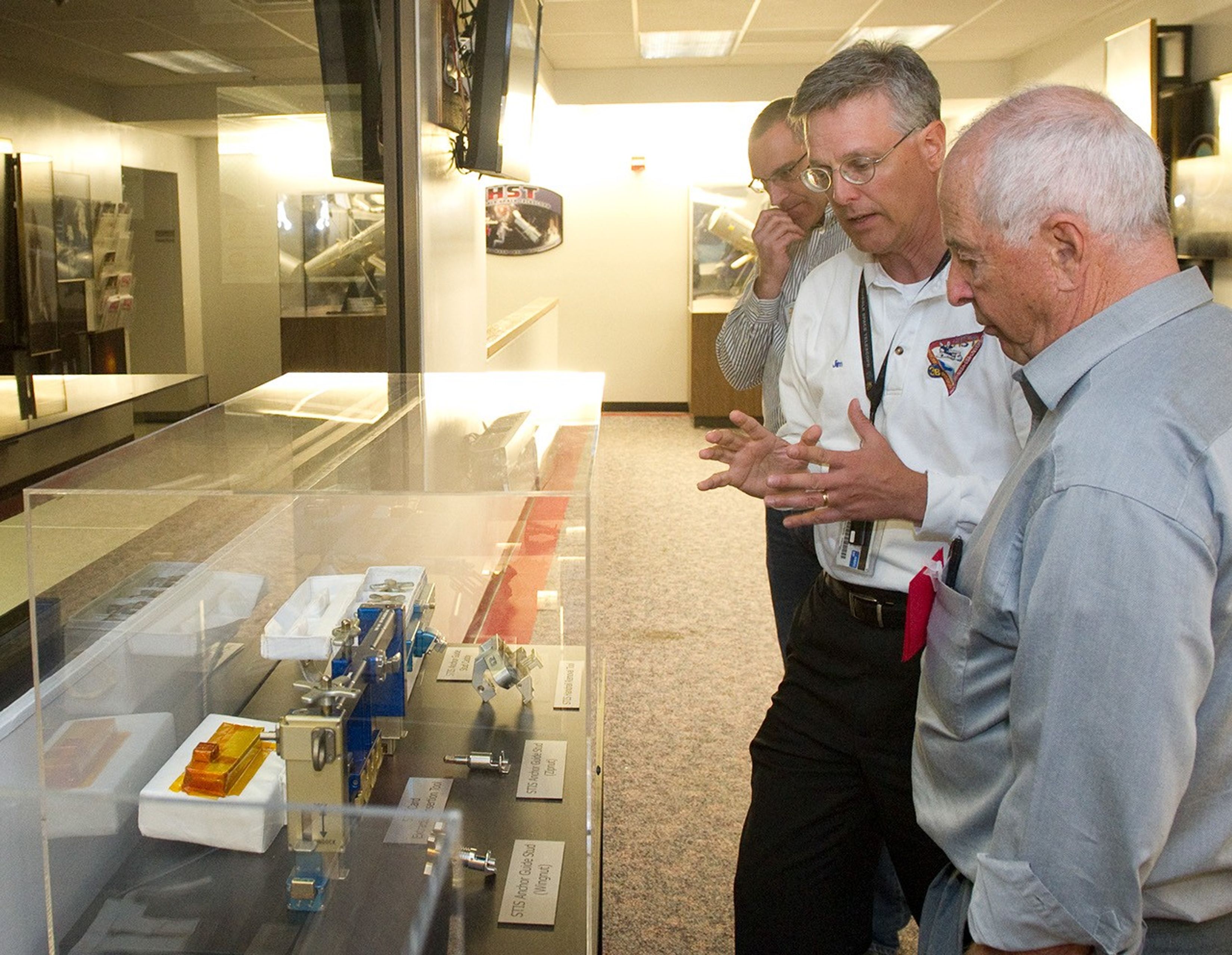 Bill Anders, Jim Jeletic, and Jim Rice stand in a line on the right side, with Anders closest to the camera, Jeletic in the middle speaking to him, and Rice on the end. They are looking at the astronaut tool case, which is on the left-hand side in the foreground.