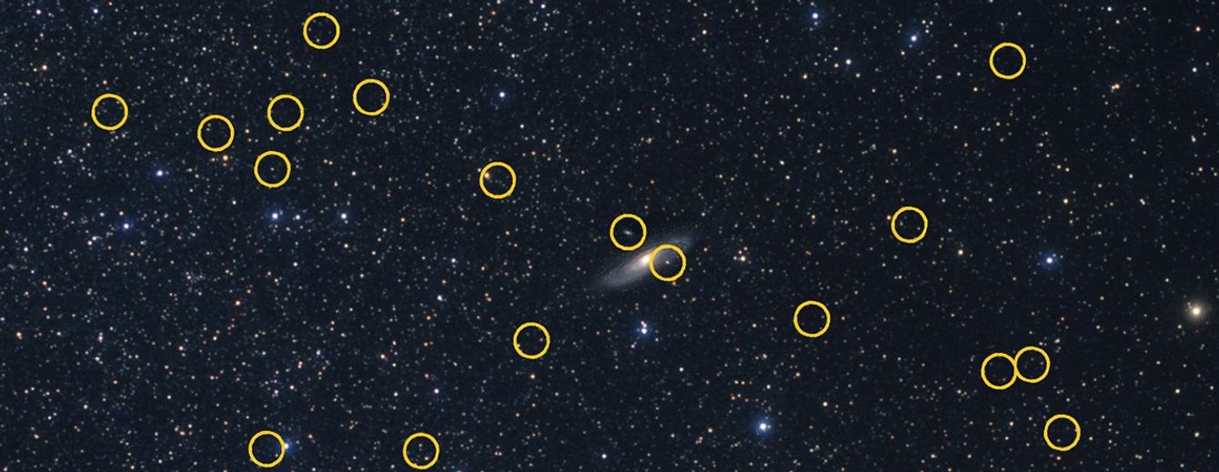 A wide-angle view of the distribution of 36 satellite galaxies orbiting the large Andromeda galaxy, which resembles bees swarming around a hive. Each dwarf is a fuzzy pinpoint identified by a yellow circle. Most of the photo is dominated by myriad pinpoint-white foreground stars inside our Milky Way galaxy.
