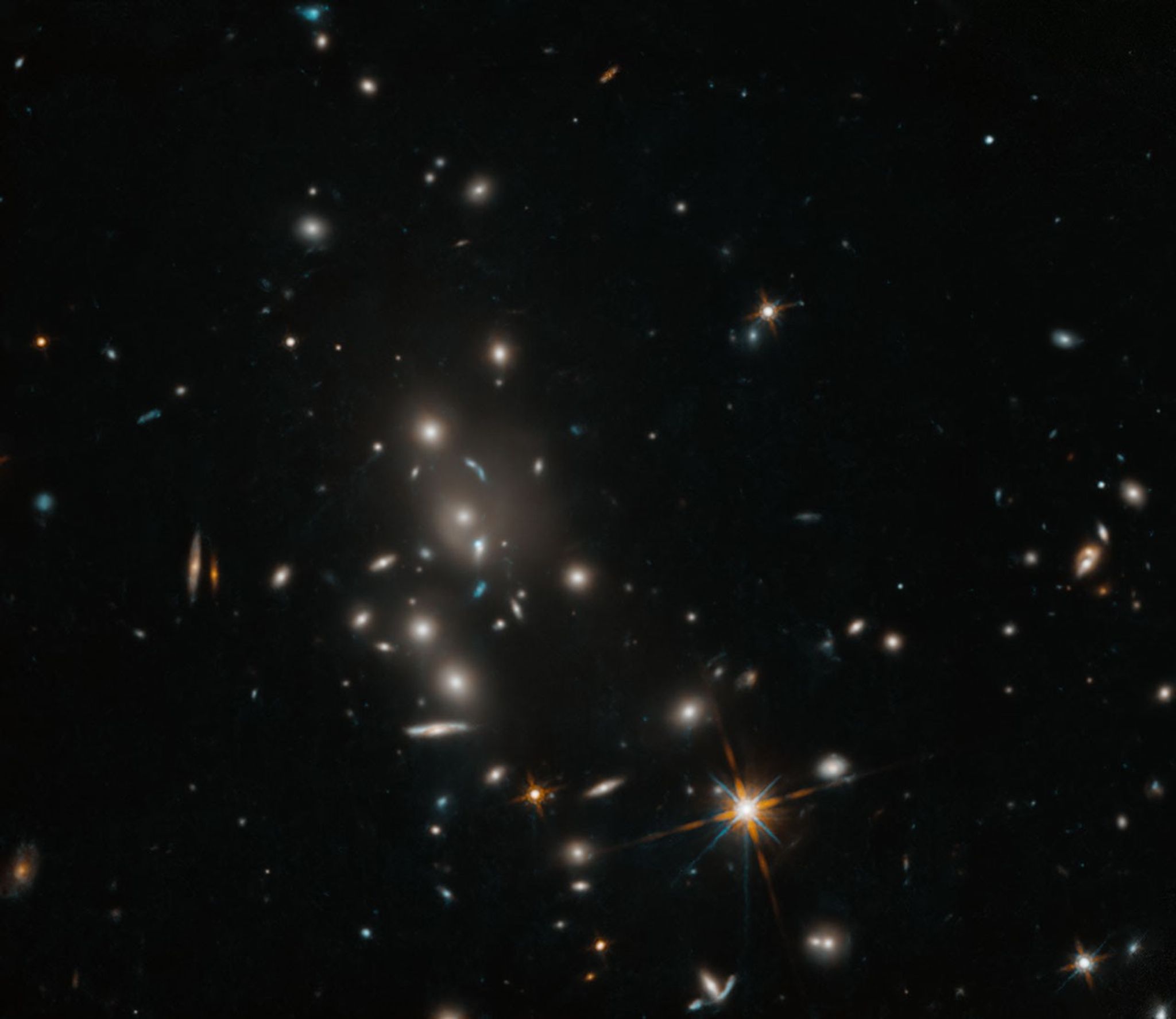 A cluster of galaxies dots the field of view. The cluster holds spiral and elliptical galaxies against a black background. A foreground star with diffraction spikes shines brightly toward the bottom, center-right of the image.