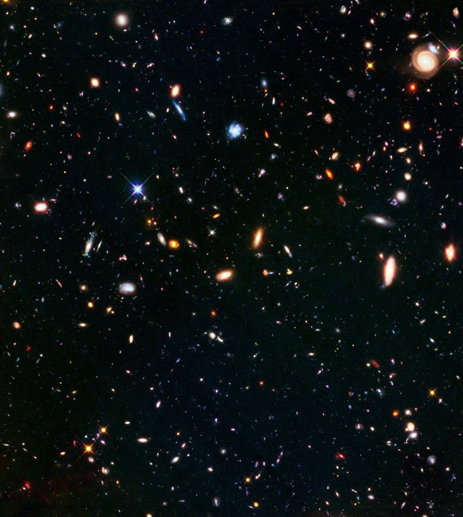 Galaxies glow in a myriad of colors against the black background of space. An orange spiral faces the camera head-on in the upper-right.