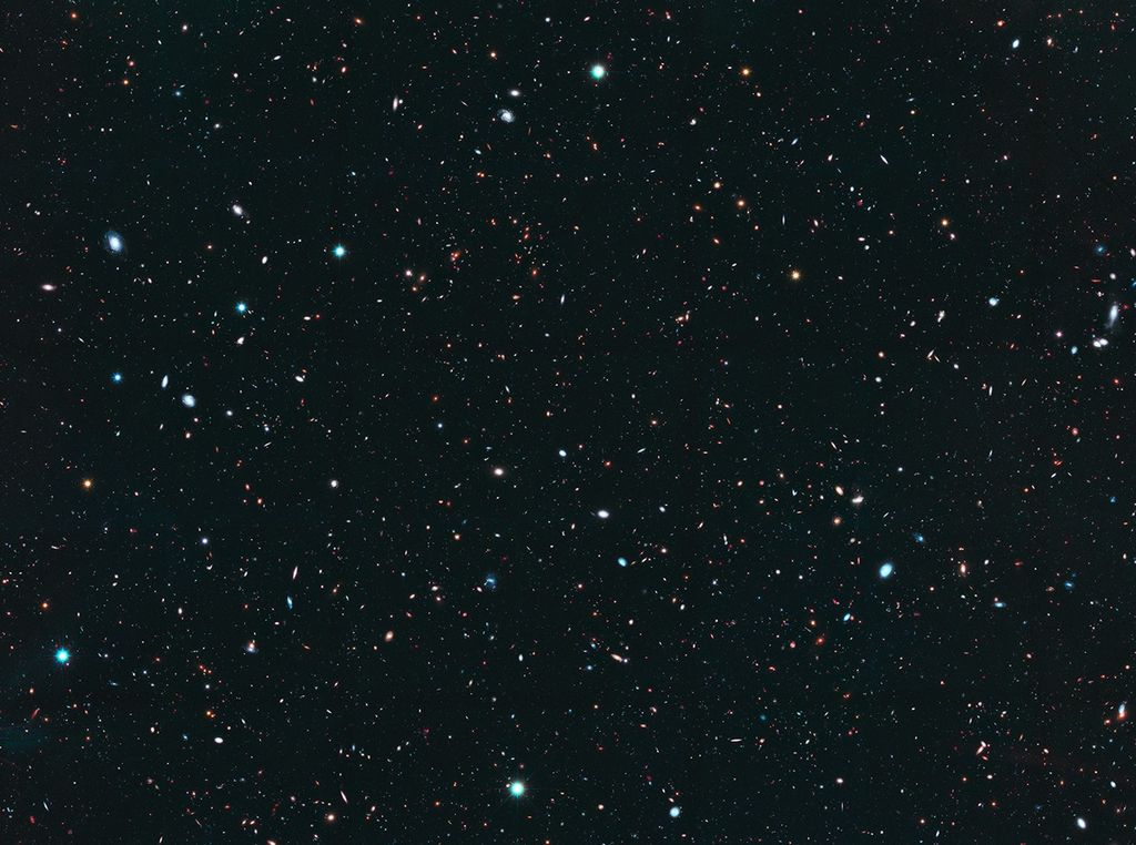 Image is filled with distant galaxies in colors of yellow-gold, red, blue, white. Many of the stars and galaxies have a bluish tint.
