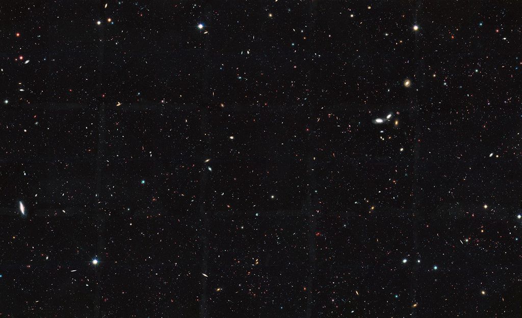 Image is filled with distant galaxies in colors of yellow-gold, red, blue, white. A clump of multi-colored galaxies that appear larger are visible on the center-right, and another spiral galaxy on the far-left.