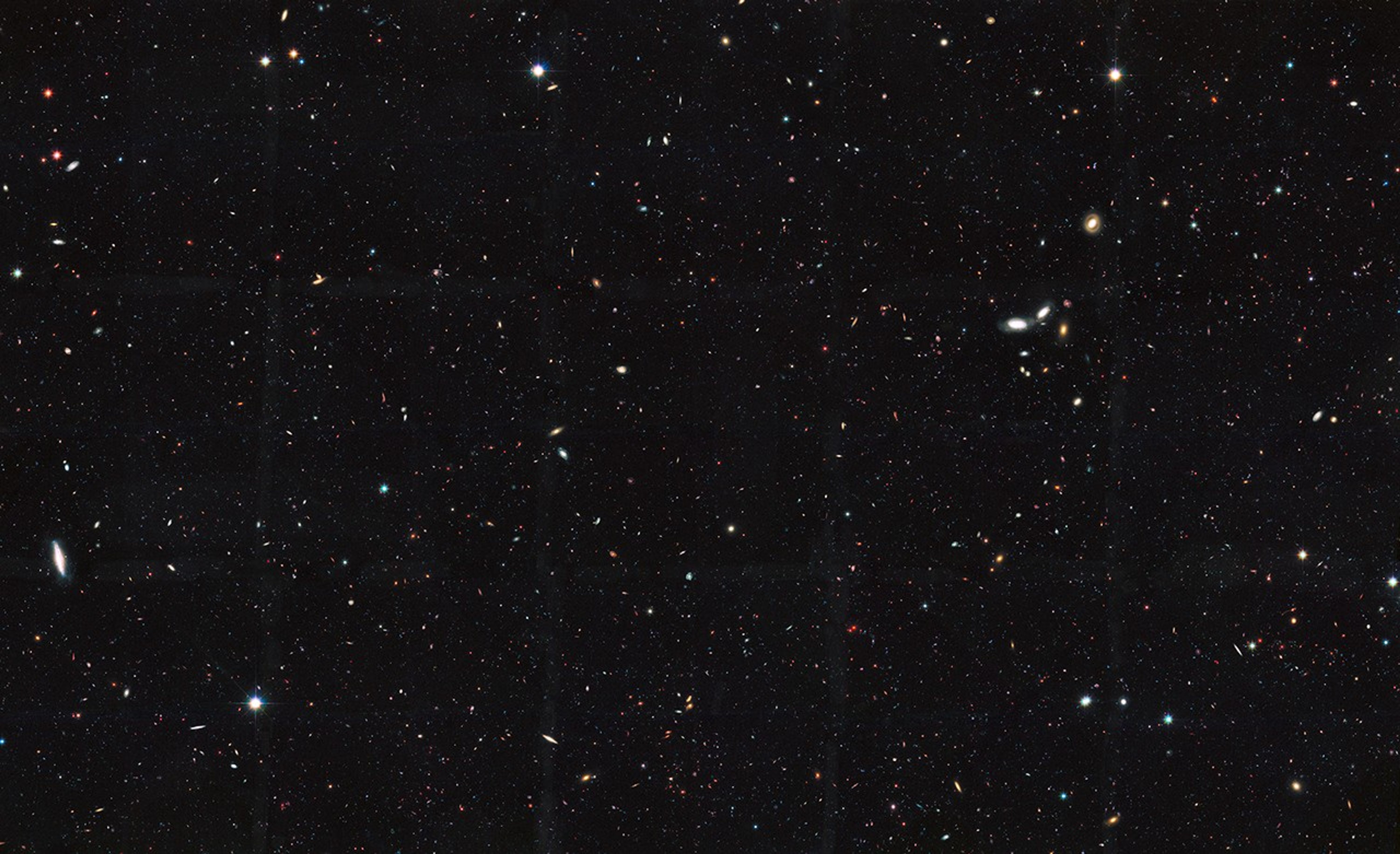 Image is filled with distant galaxies in colors of yellow-gold, red, blue, white. A clump of multi-colored galaxies that appear larger are visible on the center-right, and another spiral galaxy on the far-left.