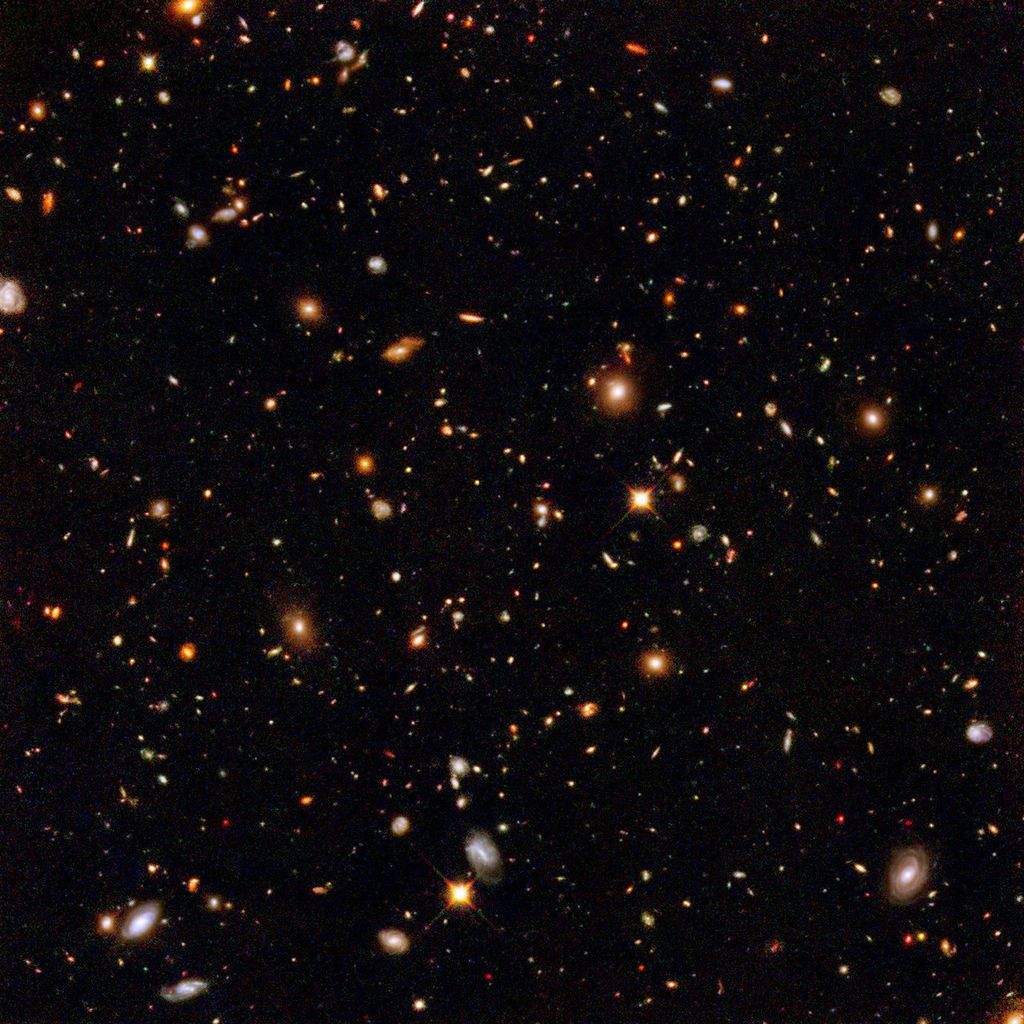 Image is filled with distant galaxies in colors of yellow-gold and reddish hues. Lower left holds a face on spiral galaxy.